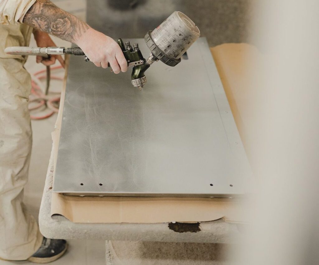 Hand holding a spray gun over a bare metal panel on a work table